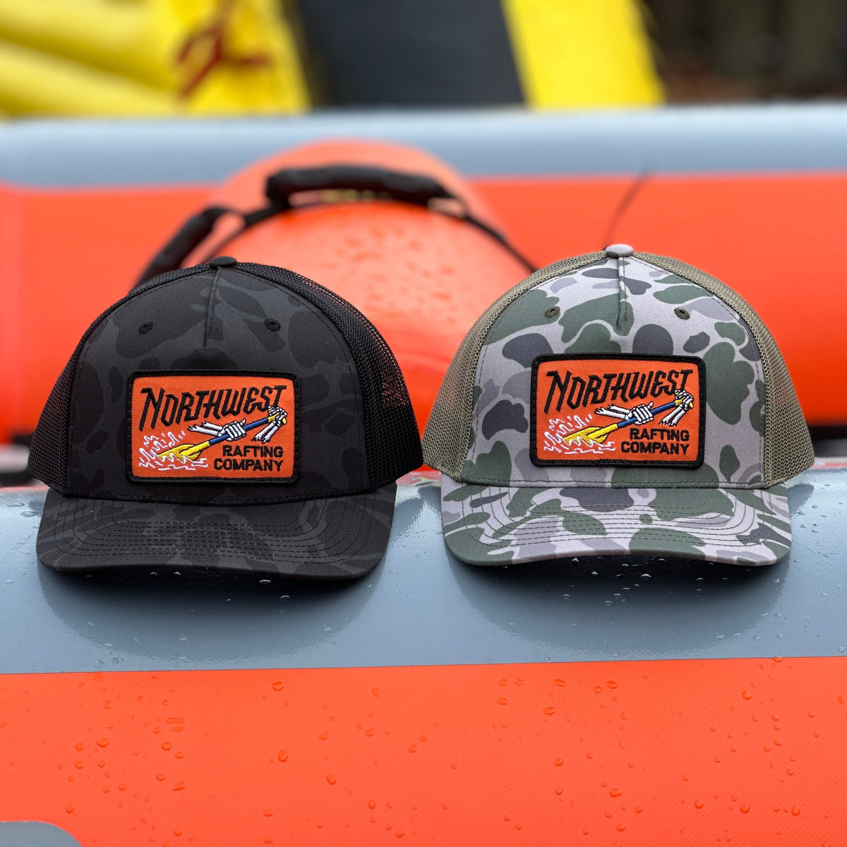 Northwest Rafting Company (NWRC) Work Hat in the Sable Duck Camo/Black (Grey/Black) and Marsh Duck Camo/Loden (Green/Tan) colors sitting on a whitewater raft